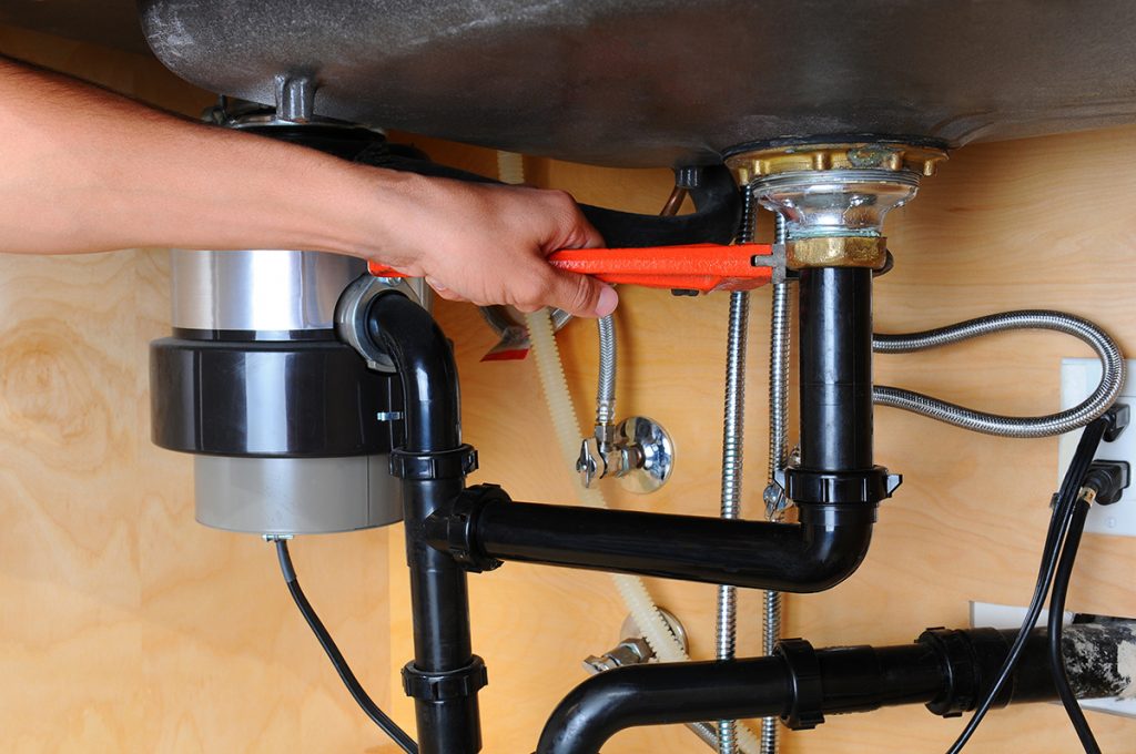 A plumber works on a garbage disposal and other pipes under a sink